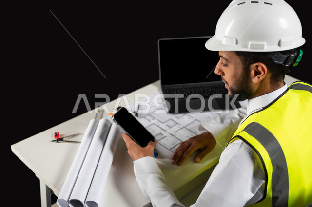 استثمار التكنولوجيا والتقنيات الحديثة في تطور المملكة العربية السعودية, بورتريه لمهندس عربي خليجي سعودي يرتدي سترة الحماية و الخوذة  البيضاء يجلس وراء المكتب ويمسك الجوال، مخططات البناء  والحاسوب المحمول على الطاولة المكتبية, خلفية سوداء
