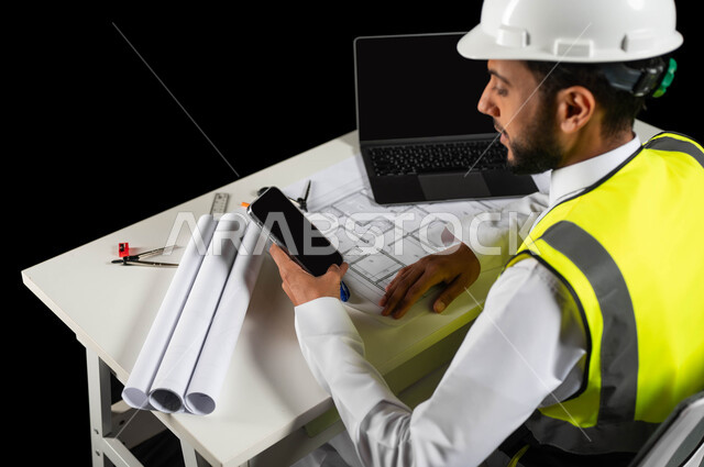 متابعة آخر مستجدات التكنولوجيا الهندسية عبر الهاتف المحمول, بورتريه لمهندس عربي خليجي سعودي يرتدي سترة الحماية و الخوذة البيضاء يجلس وراء المكتب يمسك جهاز الجوال بيده, هندسة الأبنية و الإنشاءات، مخططات البناء  والحاسوب المحمول على الطاولة المكتبية, خلفية سوداء