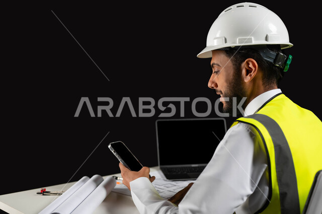 دمج التكنولوجيا والتقنية بالعمل, بورتريه مقرب من الجانب لمهندس عربي خليجي سعودي يرتدي سترة الحماية و الخوذة  البيضاء يجلس وراء المكتب, النظر إلى شاشة الجوال,  مخططات البناء والحاسوب المحمول على الطاولة المكتبية,هندسة الأبنية و الإنشاءات، خلفية سوداء