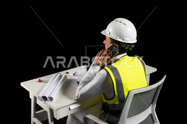 تنمية وتطوير المجالات الهندسية، بورتريه لمهندس عربي خليجي سعودي يرتدي سترة واقية وخوذة بيضاء يجلس خلف المكتب ويجري مكالمة عبر الهاتف المحمول، مخططات البناء والكمبيوتر المحمول على طاولة المكتب، الإشراف على المشاريع، هندسة البناء والإنشاءات، خلفية سوداء