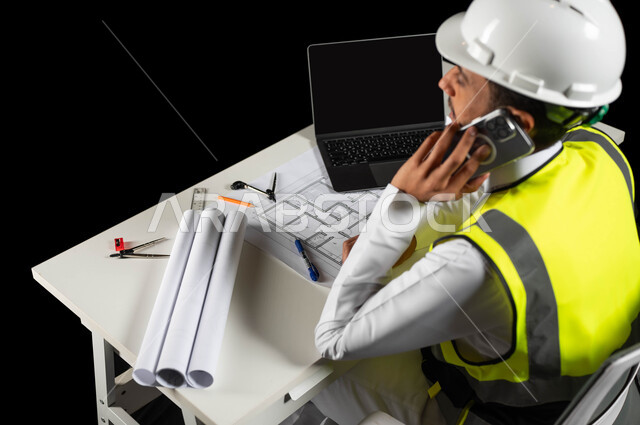 بناء و تطور المملكة العربية السعودية بأعمال أبناء الوطن, بورتريه لمهندس عربي خليجي سعودي  يرتدي سترة الحماية والخوذة  البيضاء يجلس وراء المكتب ويجري مكالمة عبر الهاتف المحمول, هندسة الأبنية و الإنشاءات، مخططات البناء واللابتوب على الطاولة المكتبية, خلفية سوداء