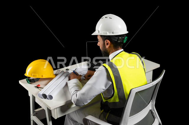 مفهوم الهندسة الإنشائية، العمل في القطاع الهندسي, بورتريه من الخلف لمهندس عربي خليجي سعودي يرتدي خوذة الوقاية البيضاء وسترة الحماية الخاصة بالعمل يجلس على المكتب, المخططات المعمارية والخوذة الصفراء وجهاز الحاسوب المحمول على طاولة مكتبية, خلفية سوداء