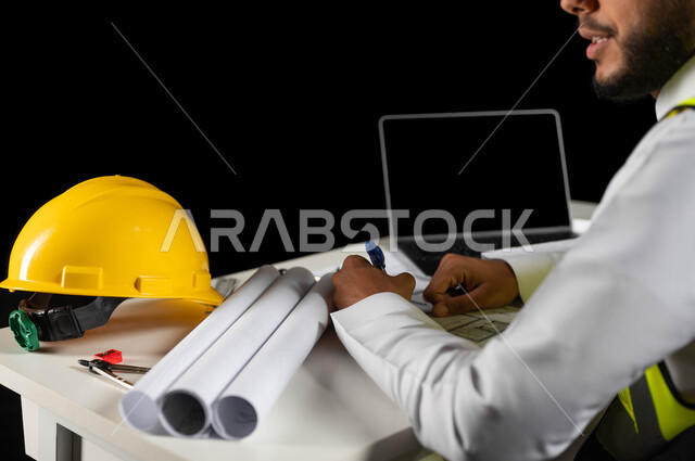 إنجاز مهام العمل ومتابعة سير المشروع، خوذة الوقاية الصفراء والحاسوب المحمول على المكتب، مهن ووظائف هندسية سعودية، الأدوات ومخططات البناء على طاولة مكتبية، بورتريه مقرب ليد مهندس عربي خليجي سعودي يقوم بتدوين بعض الملاحظات حول خطة البناء, خلفية سوداء