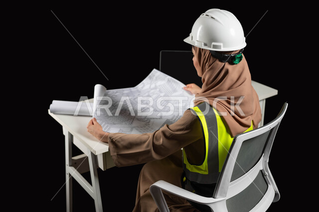 Auditing and reviewing construction plans, portrait from the side of a veiled Saudi Gulf Arab engineer wearing a protection jacket and helmet holding the construction plan and displaying it in front of her, engineering project management, women’s professions and jobs, continuing work in the office, black background