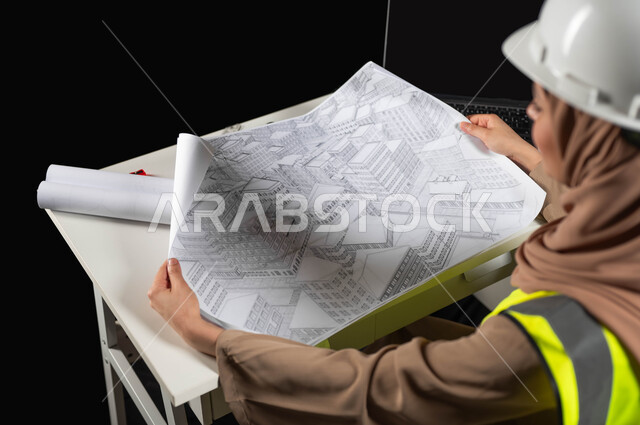 مفهوم الهندسة والانشاءات المعمارية, عمل النساء في القطاع الهندسي, بورتريه مقرب من الجانب لمهندسة عربية خليجية سعودية محجبة ترتدي سترة الحماية والخوذة تمسك مخطط الإعمار, مراجعة وعرض مخطوطات المشاريع بشكل كامل, إدارة ومتابعة المشروع, خلفية سوداء