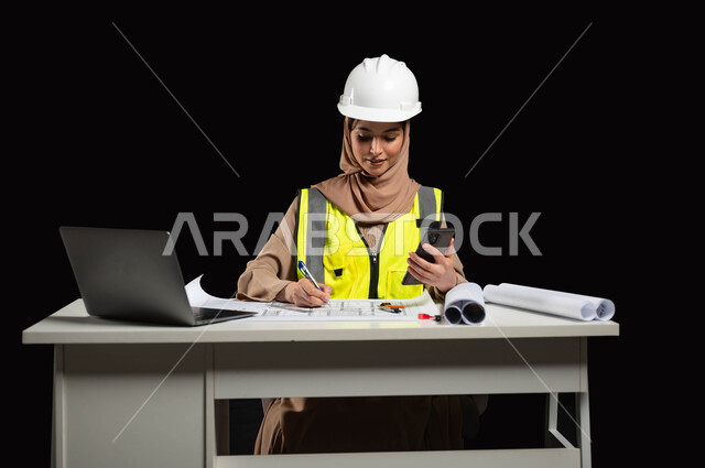 دمج التكنولوجيا الحديثة في القطاع الهندسي, متابعة المشاريع الإنشائية عبر الهاتف المحمول, بورتريه لمهندسة عربية خليجية سعودية محجبة ترتدي سترة الحماية تقوم بالتعديل على مخطط الإعمار عن طريق التطبيقات والمواقع الهندسية, إدارة ومتابعة العمل، خلفية سوداء
