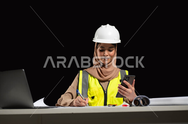 إدارة ومتابعة المشاريع الإنشائية عبر الهاتف المحمول, دمج التقنية والتكنولوجيا في المجال الهندسي, بورتريه لمهندسة عربية خليجية سعودية محجبة ترتدي الخوذة وسترة الحماية تقوم بتدوين الملاحظات, التعديل على مخطط الإعمار بواسطة التطبيقات والمواقع الهندسية, خلفية سوداء