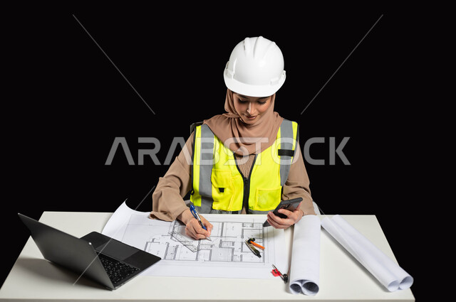 متابعة العمل عبر الهاتف المحمول,الاستفادة من التكنولوجيا الحديثة في مجال الإنشاءات المعمارية, بورتريه لمهندسة عربية خليجية سعودية محجبة ترتدي الخوذة وسترة الحماية الخاصة بالعمل, التعديل على مخطط الإعمار عن طريق التطبيقات والمواقع الهندسية, خلفية سوداء