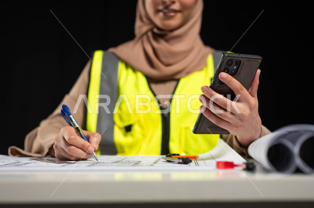 الاستفادة من التكنولوجيا الحديثة في مجال العمل, متابعة المشاريع الإنشائية عبر الهاتف المحمول, بورتريه مقرب لمهندسة عربية خليجية سعودية محجبة ترتدي سترة الحماية تمسك بيدها القلم لتقوم بالتعديل على مخطط الإعمار عن طريق التطبيقات والمواقع الهندسية, خلفية سوداء