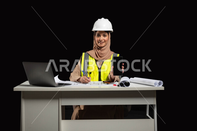 The development of modern technologies and technology in the Kingdom of Saudi Arabia, a portrait of a Saudi Arabian Gulf engineer wearing a white helmet and work jacket, sitting at the desk and displaying the mobile device with a black screen, looking at the camera with gestures of joy and happiness, black background
