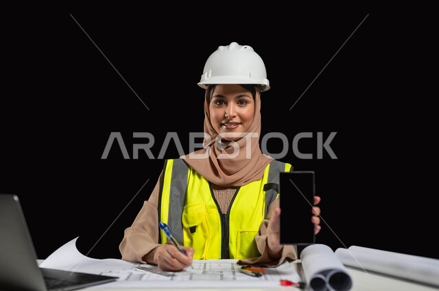 استخدام التقنيات الحديثة لتسهيل القيام بالمهام المطلوبة, دمج العمل مع التكنولوجيا، عرض شاشة جوال سوداء, بورتريه لمهندسة عربية خليجية سعودية ترتدي الخوذة البيضاء والسترة الخاصة بالعمل تجلس على المكتب، النظر إلى الكاميرا بإيماءات السرور, خلفية باللون الأسود