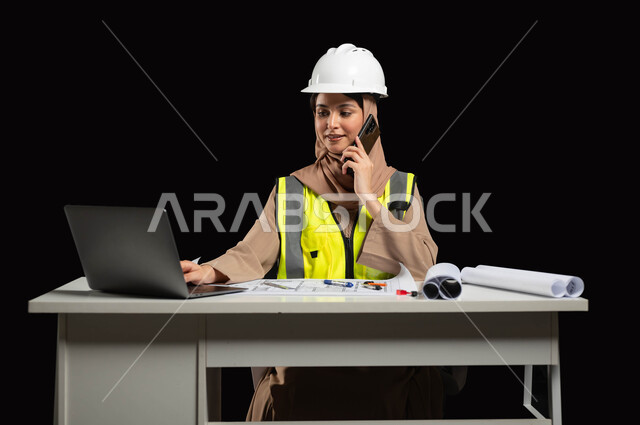 Browsing engineering websites and applications, integrating work with technology and technology, portrait of a Saudi Arabian Gulf engineer wearing a white helmet and work jacket sitting at the desk, making a call via mobile phone and reviewing the latest developments via the laptop, black background