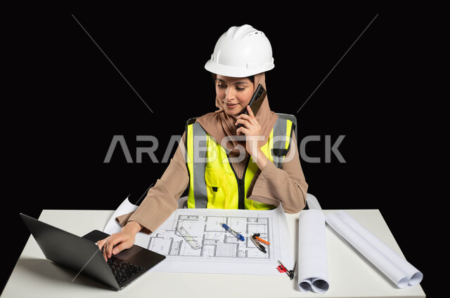 دمج العمل مع التقنية و التكنولوجيا، الاطلاع على تصاميم هندسية جديدة, بورتريه لمهندسة عربية خليجية سعودية ترتدي الخوذة البيضاء والسترة الخاصة بالعمل تجلس على المكتب، إجراء مكالمة عبر الهاتف المحمول ومطالعة آخر التطورات عن طريق جهاز اللابتوب, خلفية سوداء