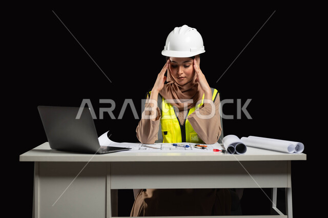 الإشراف على المشاريع الهندسية, الجد والاجتهاد في العمل, بورتريه لمهندسة عربية خليجية سعودية محجبة ترتدي الخوذة وسترة الحماية تمسك رأسها بيديها بإيماءات تدل على الشعور بالصداع, التعب والإرهاق والحاجة إلى الراحة، مساهمة المرأة في المجتمع، خلفية سوداء