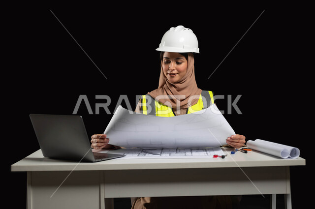 مراجعة المخططات الهندسية, الإشراف على سير العمل, بورتريه لمهندسة عربية خليجية سعودية محجبة  ترتدي خوذة الحماية والسترة الخاصة بالعمل وتجلس على المكتب وتمسك بيدها مخطط الإنشاء،عمل المرأة السعودية في القطاع الهندسي، خلفية سوداء