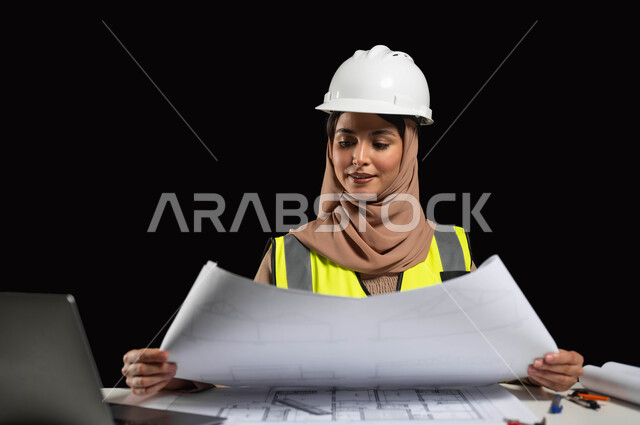 مفهوم الهندسة والإنشاءات المعمارية، بورتريه مقرب لمهندسة عربية خليجية سعودية محجبة ترتدي خوذة الحماية والسترة الخاصة بالعمل وتجلس على المكتب وتمسك بيدها مخطط هندسي، إيماءات الفخر بالوصول إلى حلم النجاح، الإشراف على العمل، خلفية سوداء