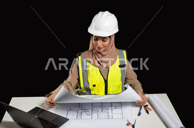 Professions and jobs for Saudi women, the concept of engineering and architectural construction, a portrait of a veiled Saudi Gulf Arab engineer wearing a protective helmet and work jacket, sitting at the desk and holding engineering plans in her hand, supervising the work, black background