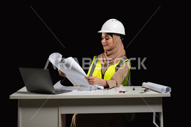 استخدام الحاسوب المحمول في القطاع الهندسي، النظر الى المخطط الهندسي بتعابير السرور، مفهوم ادارة المشاريع وتدقيقها، بورتريه مقرب لمهندسة عربية خليجية سعودية محجبة ترتدي سترة الحماية والخوذة  تقوم بمراجعة المخططات الإنشائية، خلفية سوداء, محتوى حساس، استخدام حساس