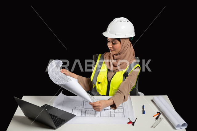 النظر الى المخطط الهندسي بتعابير السرور، العمل في القطاع الهندسي، مفهوم ادارة المشاريع وتدقيقها، بورتريه مقرب لمهندسة عربية خليجية سعودية محجبة ترتدي سترة الحماية والخوذة  تقوم بتدقيق ومراجعة المخططات الإنشائية، خلفية سوداء, محتوى حساس، استخدام حساس