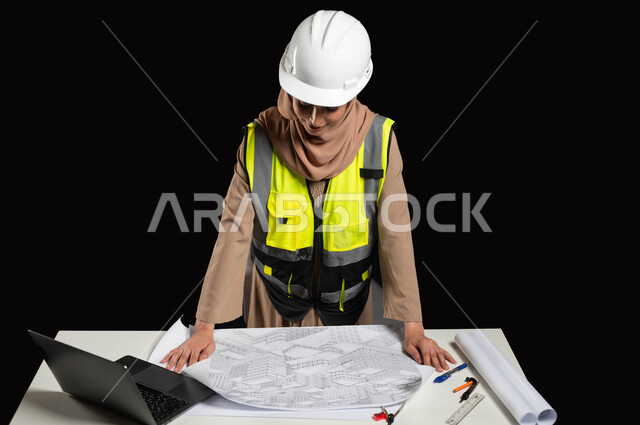 العمل في القطاع الهندسي، دور الهندسة المعمارية في تطور المجتمع السعودي، مفهوم ادارة المشاريع وتدقيقها، بورتريه مقرب لمهندسة عربية خليجية سعودية محجبة ترتدي سترة الحماية والخوذة  تقوم بتدقيق ومراجعة المخططات الإنشائية، خلفية سوداء,محتوى حساس، استخدام حساس
