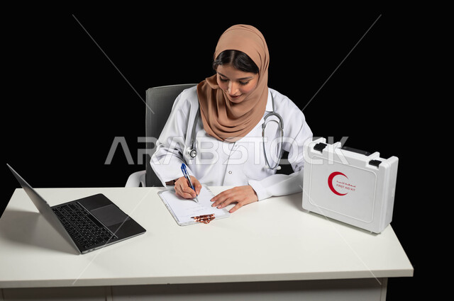 كتابة وتدوين التقارير والحالات الصحيه، تقديم الخدمات الطبية للمرضى و متابعة حالاتهم, استخدام حقيبة الاسعافات الاوليه،  بورتريه مقرب لطبيبة عربية خليجية ترتدي المعطف الأبيض تقوم باستعمال الحاسوب المحمول، خلفية سوداء, محتوى حساس, استخدام حساس