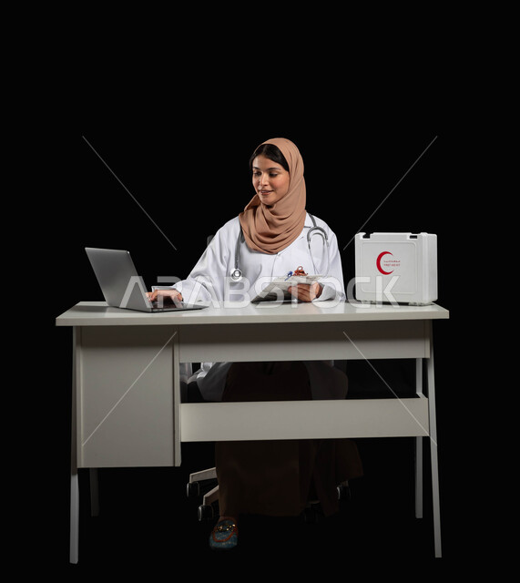 دمج العمل الطبي مع التكنولوجيا، استخدام حقيبة الاسعافات الاوليه والحاسوب المحمول في مجال الطب، بورتريه مقرب لطبيبة عربية خليجية ترتدي المعطف الأبيض تقوم بكتابة وتدوين التقارير والحالات الصحية، خلفية سوداء, محتوى حساس, استخدام حساس