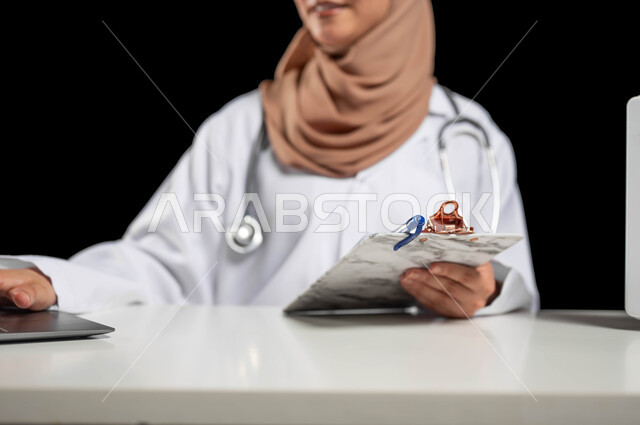 مفهوم الطب والرعاية الصحية، استخدام حقيبة الاسعافات الاوليه والحاسوب المحمول في مجال الطب، دمج العمل مع التكنولوجيا, بورتريه مقرب لطبيبة عربية خليجية ترتدي المعطف الأبيض تقوم بكتابة وتدوين التقارير والحالات الطبية، خلفية سوداء, محتوى حساس, استخدام حساس