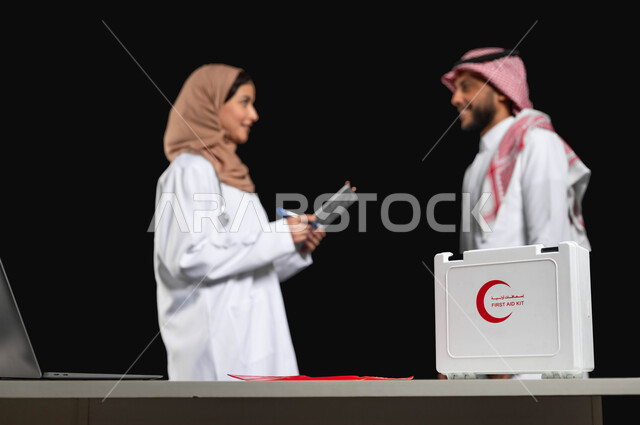 الرعاية الطبية في السعودية، ايماءات البشاشه في التعامل مع المرضى، بورتريه مقرب لطبيبة عربية خليجية سعودية محجبة تشرح الحاله الصحيه لمريض يرتدي الثوب والشماغ التقليدي، استخدام حقيبة الإسعافات الاوليه وجهاز الحاسوب في الطب, خلفية سوداء, محتوى حساس, استخدام حساس