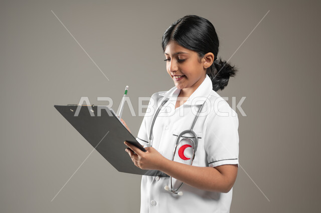 طموحات الأطفال في العمل في المجال الطبي، تدوين وتسجيل معلومات وبيانات المرضى، تحديد مهنة المستقبل منذ الصغر، بورتريه لطفلة عربية خليجية سعودية ترتدي زي ممرضة تمسك بيدها ملف ملاحظات، خلفية رمادية، محتوى حساس، استخدام حساس