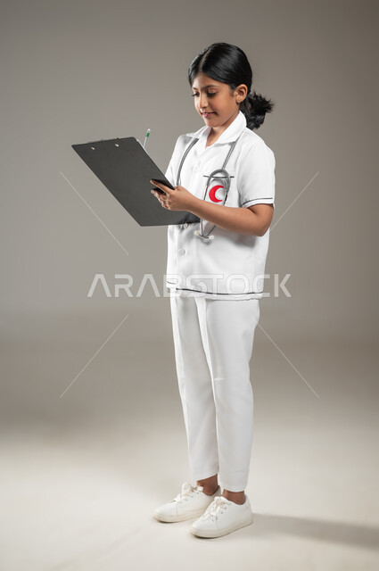 تدوين وتسجيل معلومات وبيانات المرضى، تحديد مهنة المستقبل منذ الصغر، بورتريه لطفلة عربية خليجية سعودية ترتدي زي ممرضة تمسك بيدها ملف ملاحظات، صورة لكامل الجسم، خلفية رمادية، محتوى حساس، استخدام حساس