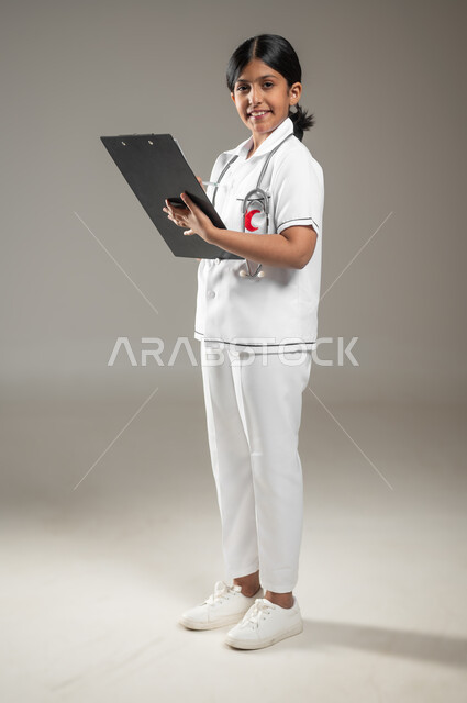 Recording patient information and data, determining the future profession since childhood, portrait of a Saudi Gulf Arab girl wearing a nurse’s uniform, holding a file of notes in her hand, looking at the camera with gestures of joy and pleasure, full-body photo, gray background, sensitive content, sensitive use