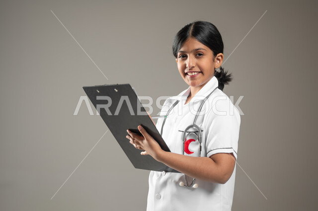 تحديد مهنة المستقبل منذ الصغر، بورتريه لطفلة عربية خليجية سعودية ترتدي زي ممرضة يحمل شعار الهلال الأحمر السعودي تمسك بيدها ملف ملاحظات تنظر إلى الكاميرا بإيماءات البهجة والسرور، تسجيل معلومات وبيانات المرضى، خلفية رمادية، محتوى حساس، استخدام حساس