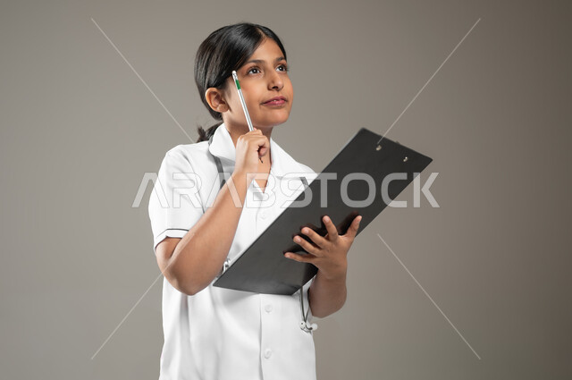 ايماءات التركيز والتفكير بشيء ما، تدوين وتسجيل معلومات وبيانات المرضى، تحديد مهنة المستقبل منذ الصغر، بورتريه مقرب لطفلة عربية خليجية سعودية ترتدي زي ممرضة تمسك بيدها ملف ملاحظات، خلفية رمادية، محتوى حساس، استخدام حساس