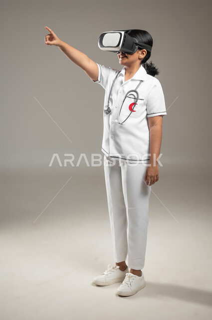 الوقوف باستقامة والإشارة الى شيء ما، دمج التكنولوجيا الحديثة في المجال الطبي، تحديد مهنة المستقبل منذ الصغر، بورتريه لطفلة عربية خليجية سعودية ترتدي زي الممرضة تضع نظارة العالم الافتراضي، صورة لكامل الجسم، خلفية رمادية، محتوى حساس، استخدام حساس