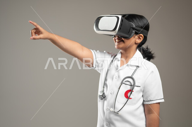 الإشارة الى شيء ما، دمج التكنولوجيا الحديثة في المجال الطبي، تحديد مهنة المستقبل منذ الصغر، بورتريه مقرب لطفلة عربية خليجية سعودية ترتدي زي الممرضة تضع نظارة العالم الافتراضي، خلفية رمادية، محتوى حساس، استخدام حساس