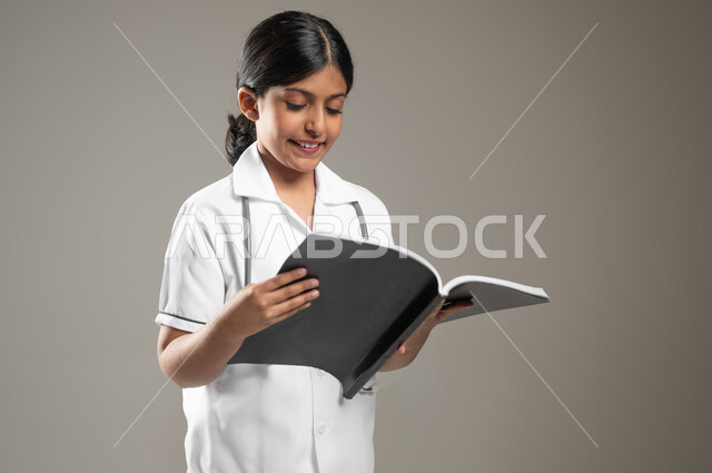 تسجيل وتدوين ملاحظات عن حالة المرضى، طموحات الأطفال في العمل في المجال الطبي، تحديد مهنة المستقبل منذ الصغر، بورتريه مقرب لطفلة عربية خليجية سعودية ترتدي لباس الممرضة تقوم بمراجعة سجل المعلومات، خلفية رمادية، محتوى حساس، استخدام حساس