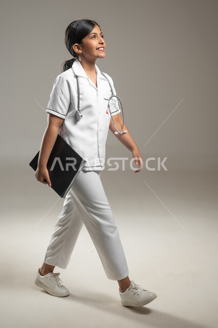 السير إلى مكان ما بإبماءات الثقة والسرور، طموحات الأطفال في العمل في المجال الطبي، تحديد مهنة المستقبل منذ الصغر، بورتريه مقرب لطفلة عربية خليجية سعودية ترتدي لباس الممرضة تحمل سجل المعلومات الخاص بالمرضى، خلفية رمادية، صورة لكامل الجسم، محتوى حساس، استخدام حساس