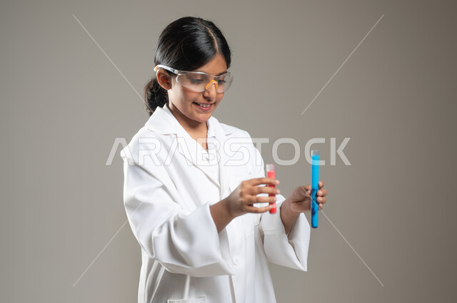 تحديد مهنة المستقبل منذ الصغر، التحاليل الطبية في المختبرات، مراقبة التفاعلات الكيميائية المجراة باستعمال أنابيب الاختبار، بورتريه مقرب لفتاة عربية خليجية سعودية ترتدي المعطف الطبي ونظارات الحماية، خلفية رمادية، محتوى حساس، استخدام حساس