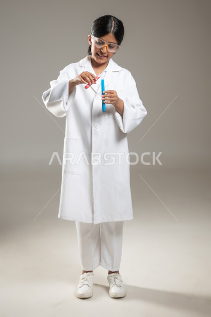 تحديد مهنة المستقبل منذ الصغر، بورتريه لفتاة عربية خليجية سعودية ترتدي المعطف الطبي ونظارات الحماية تقوم بإجراء التفاعلات الكيميائية باستخدام أنابيب الاحتبار، التحاليل الطبية في المختبرات، صورة لكامل الجسم، خلفية رمادية، محتوى حساس، استخدام حساس