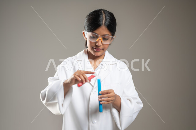 التحاليل الطبية في المختبرات، تحديد مهنة المستقبل منذ الصغر، بورتريه مقرب لفتاة عربية خليجية سعودية ترتدي المعطف الطبي ونظارات الحماية تقوم بإجراء التفاعلات الكيميائية باستخدام أنابيب الاحتبار، تجارب وأبحاث علمية مخبرية، خلفية رمادية، محتوى حساس، استخدام حساس