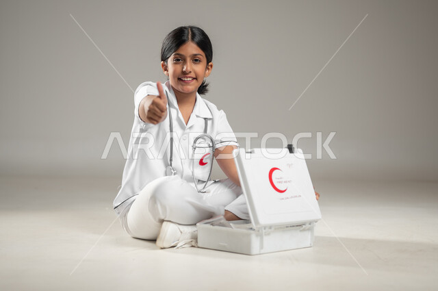 Children's ambition to be workers in the medical field, first aid kit bag, portrait of a Saudi Arabian Gulf girl wearing a nurse's uniform sitting on the floor raising her hand with a gesture of acceptance, future professions and jobs, full-body portrait, gray background, sensitive content, sensitive use