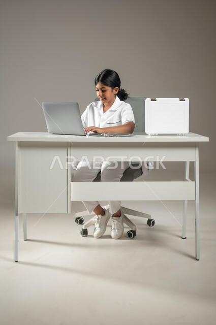 العمل في المجال الطبي، حقيبة الإسعافات الأولية، مهن ووظائف مستقبلية، استعمال التكنولوجيا الحديثة، بورتريه لطفلة عربية سعودية خليجية ترتدي زي الممرضة بالنقر على الحاسوب المحمول، صورة لكامل الجسم، خلفية رمادية، محتوى حساس، استخدام حساس