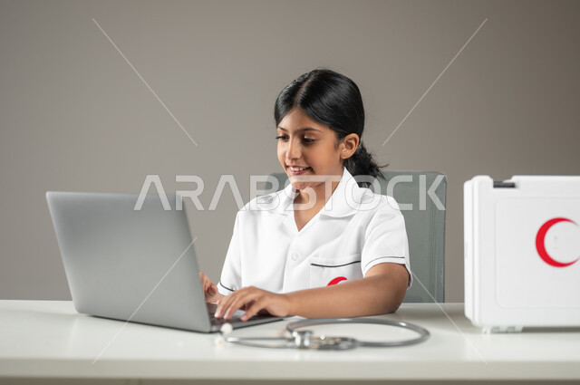 مهن ووظائف مستقبلية، دمج التكنولوجيا الحديثة في المجال الطبي، حقيبة الإسعافات الأولية، بورتريه مقرب لطفلة عربية سعودية خليجية ترتدي زي الممرضة بالنقر على الحاسوب المحمول، خلفية رمادية، محتوى حساس، استخدام حساس