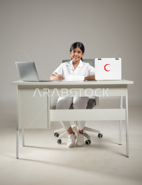 النظر إلى الكاميرا بإيماءات البهجة والسرور، استعمال التكنولوجيا في المجال الطبي، حقيبة الإسعافات الأولية، بورتريه لطفلة عربية سعودية خليجية ترتدي زي الممرضة تقوم بتدوين ملاحظات المرضى، مهن ووظائف مستقبلية، صورة لكامل الجسم، خلفية رمادية، محتوى حساس، استخدام حساس