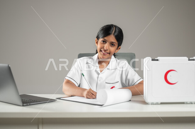 تدوين ملاحظات المرضى، استعمال التكنولوجيا الحديثة في المجال الطبي، بورتريه مقرب لطفلة عربية سعودية خليجية ترتدي زي الممرضة تجلس على مكتب، حقيبة الإسعافات الأولية، مهن ووظائف مستقبلية، خلفية رمادية، محتوى حساس، استخدام حساس