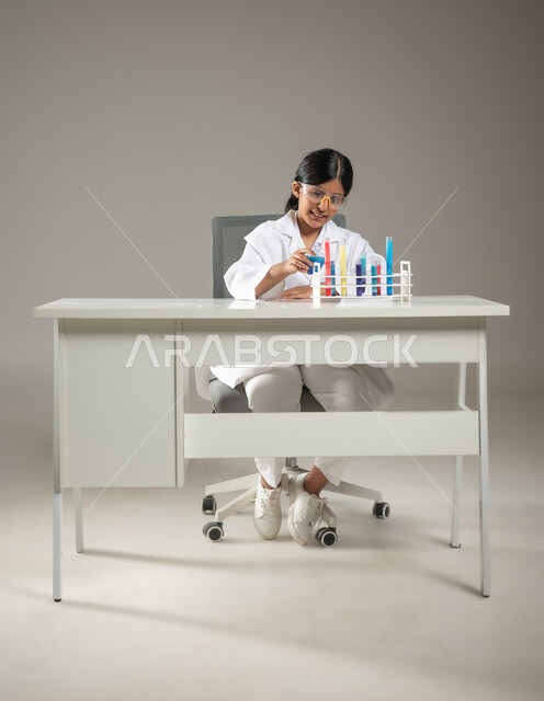 مهن ووظائف مستقبلية، تجارب وأبحاث علمية مخبرية، إجراء التحاليل الطبية، بورتريه مقرب لطفلة عربية خليجية سعودية مبتسمة ترتدي المعطف الطبي ونظارات الحماية تقوم بتفحص أنابيب إختبار التفاعلات الكيميائية، صورة لكامل الجسم، خلفية رمادية، محتوى حساس، استخدام حساس