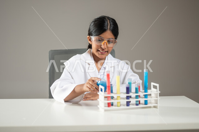 تجارب وأبحاث علمية مخبرية، إجراء التحاليل الطبية، بورتريه مقرب لطفلة عربية خليجية سعودية مبتسمة ترتدي المعطف الطبي ونظارات الحماية تقوم بتفحص أنابيب إختبار التفاعلات الكيميائية، مهن ووظائف مستقبلية ، خلفية رمادية، محتوى حساس، استخدام حساس