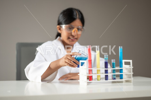 تجارب وأبحاث علمية مخبرية، إجراء التحاليل الطبية، بورتريه مقرب لطفلة عربية خليجية سعودية مبتسمة ترتدي المعطف الطبي ونظارات الحماية تقوم بإجراء التفاعلات الكيميائية باستعمال أنابيب الاختبار، مهن ووظائف مستقبلية ، خلفية رمادية، محتوى حساس، استخدام حساس