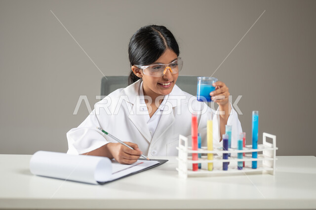 العمل في مخابر التحاليل الطبية، مراقبة التفاعلات وتدوين الملاحظات، مهن ووظائف مستقبلية، بورتريه مقرب لطفلة عربية خليجية سعودية ترتدي المعطف الطبي ونظارات الحماية تقوم بإجراء تجارب كيميائية، خلفية رمادية، محتوى حساس، استخدام حساس