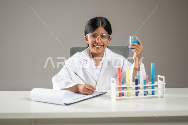 النظر إلى الكاميرا بإيماءات البهجة والسرور، تدوين وتسجيل الملاحظات، مهن ووظائف مستقبلية، بورتريه مقرب لطفلة عربية خليجية سعودية ترتدي المعطف الطبي ونظارات الحماية تقوم بإجراء تجارب كيميائية، العمل في مخابر التحاليل الطبية، خلفية رمادية، محتوى حساس، استخدام حساس